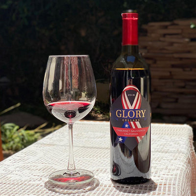 A bottle of Glory Cellars 2018 Cabernet Sauvignon next to a filled glass on a narrow table with a white cloth