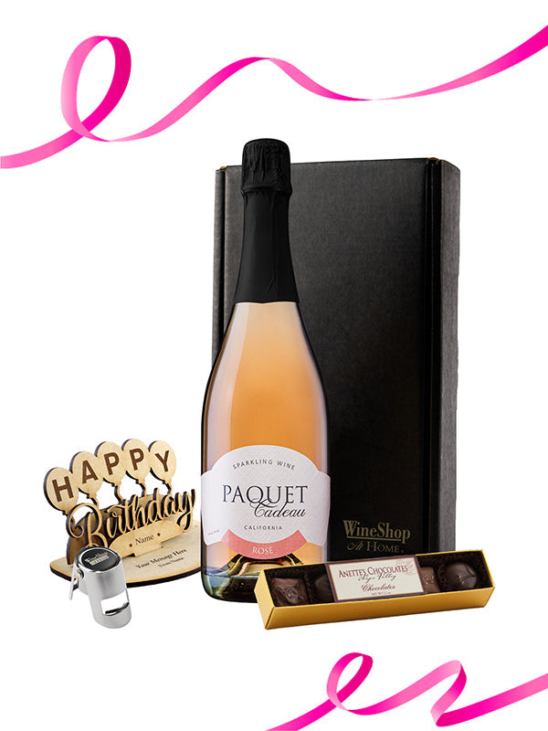 Bottle of Paquet Cuvéeau sparkling wine with a box of chocolates and a 'Happy Birthday' candle on a white background.