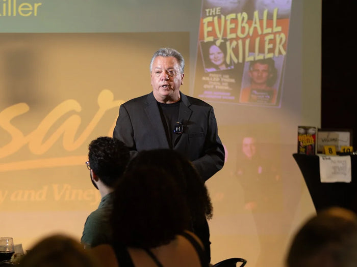 Man speaking on stage with a book titled 'The Eyeball Killer' displayed behind him