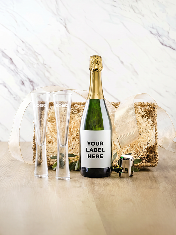 A champagne bottle with a blank white label reading 'YOUR LABEL HERE' is styled with two clear champagne flutes, a small corkscrew, and a gift box filled with shredded kraft paper and ribbon on a light wood surface against a marble-patterned backdrop.