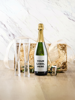 A champagne bottle with a blank white label reading 'YOUR LABEL HERE' is styled with two clear champagne flutes, a small corkscrew, and a gift box filled with shredded kraft paper and ribbon on a light wood surface against a marble-patterned backdrop.