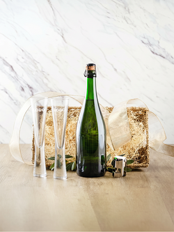 A green glass bottle with a cork closure stands on a light wood surface in front of a woven gift basket lined with straw and tied with a cream ribbon. Two clear champagne flutes and a small metal corkscrew are placed beside it against a white marble-look background.