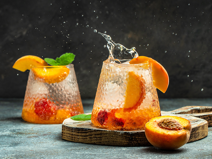 Two glasses of peach-themed cocktails with peaches and mint leaves on a dark background